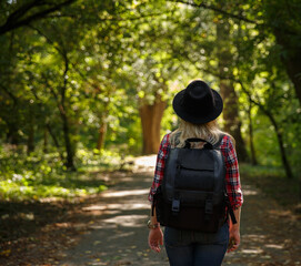 Fototapeta na wymiar young caucasian girl travels in a hat and with a backpack through the forest or park in the shade of trees .background blurred. 