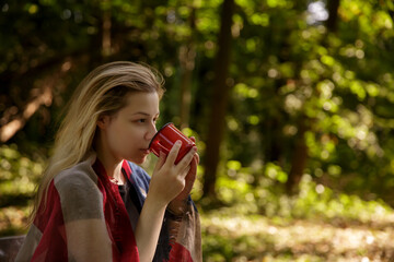 young caucasian woman drinking from red metal mug in nature.background blurred. selective focus. 