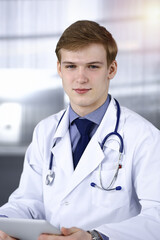 Blond male doctor sitting and working with tablet computer in sunny clinic at his working place, close-up. Medicine concept