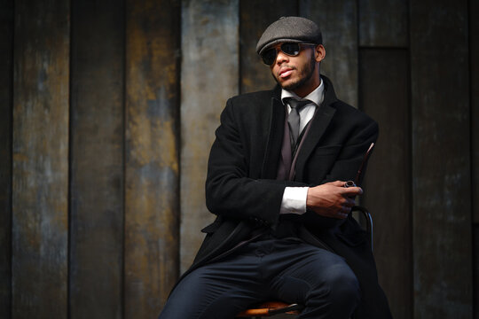 Studio shot of stylish african american man wearing coat, cap, glasses and holding smoking pipe