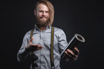 A bearded stylish man in a wheelhouse with suspenders smiles looking at the camera and holds a stainless steel muffler pointing at it with his hand. Ideal seller of car spare parts © yanik88