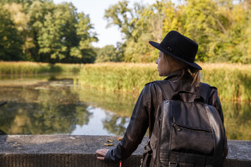 a young Caucasian woman in a leather jacket and hat and with a backpack stands by the pond and looks into the distance. selective focus. High quality photo