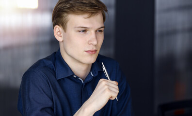 Young blond businessman thinking about strategy at his working place with computer in a darkened office, glare of light on the background