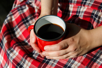 hands hold a red metal mug with a hot drink on a checkered background. selective focus. High...