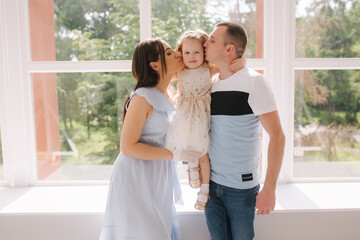 Family portrait of mom dand and little daughter in front of big window. Happy family. Mother and father kiss daughter in cheek