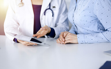 Unknown woman-doctor is showing to her patient a description of medication, while sitting together at the desk in the sunny cabinet in a clinic. Female physician is using a computer tablet and a