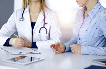 Unknown woman- doctor is listening to her patient, while sitting together at the desk in the sunny cabinet in a clinic. Female physician with a stethoscope is writing at clipboard, close up. Perfect