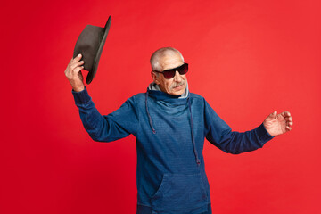 Greeting. Portrait of senior man in stylish eyewear and hat isolated on red studio background. Tech and joyful elderly lifestyle concept. Trendy colors, forever youth. Copyspace for your ad.