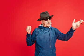 Pointing, smoking. Portrait of senior man in stylish eyewear and hat isolated on red studio...