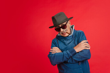 Smiling, smoking. Portrait of senior man in stylish eyewear and hat isolated on red studio background. Tech and joyful elderly lifestyle concept. Trendy colors, forever youth. Copyspace for your ad.