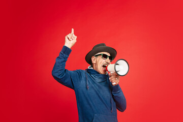 Shouting with megaphone. Portrait of senior man in stylish eyewear, hat isolated on red studio...