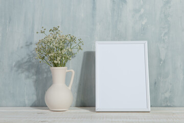 A vase with flowers and a white photo frame on the table. Light stylish background. Preparation for inscriptions, copy space.