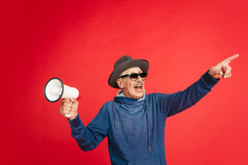 Shouting with megaphone. Portrait of senior man in stylish eyewear, hat isolated on red studio...