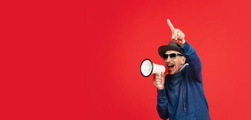 Shouting with megaphone. Portrait of senior man in stylish eyewear and hat isolated on red studio...
