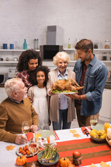 Selective focus of elderly woman holing turkey near multiethnic family during thanksgiving dinner