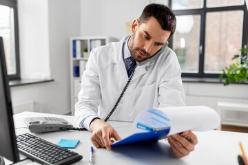 Fototapeta premium healthcare, medicine and people concept - male doctor with clipboard calling on desk phone at hospital
