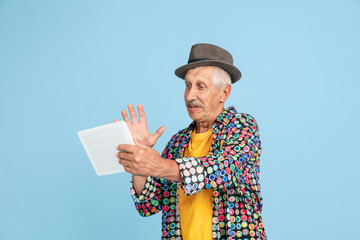 Videochat using tablet. Portrait of senior hipster man in stylish hat isolated on blue studio...