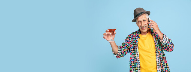 Drinks cocktail, talking phone. Portrait of senior hipster man in stylish hat on blue studio...