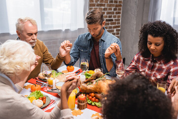 selective focus of multiethnic family with closed eyes holding hands during dinner on thanksgiving...