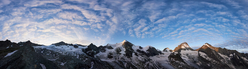 Panoramic snapshot of a ridge of rocky mountains in Switzerland in the morning, blue sky with wavy clouds is over snowy peaks, rising sun shining at Ober Gabelhorn and Dent Blanche