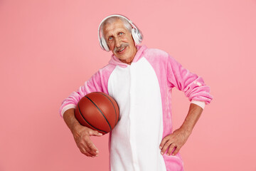 Posing with ball and headphones. Portrait of senior hipster man in stylish attire isolated on pink...