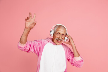Pointing up in headphones. Portrait of senior hipster man in stylish attire isolated on pink studio background. Tech and joyful elderly lifestyle concept. Trendy colors, forever youth. Copyspace © master1305