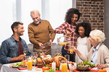joyful african american woman and senior man pouring orange juice and white wine during thanksgiving dinner with multiethnic family