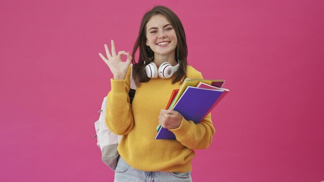A lucky student woman with headphones is showing ok gesture standing isolated over a pink background