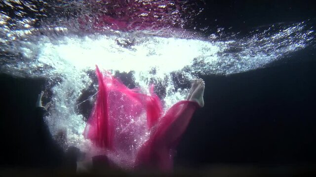 beautiful dressed woman is plunging inside water, underwater shot of her body and bubbles in light