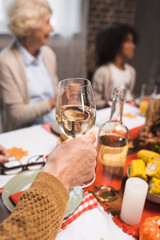 senior man holding glass of white wine while celebrating thanksgiving with multicultural family