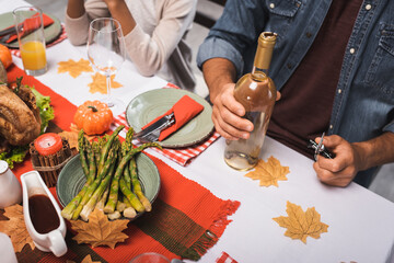 cropped view of man holding bottle of wine while sitting near african american girl during thanksgiving dinner