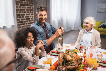selective focus of man opening bottle of white wine during thanksgiving dinner with multicultural family