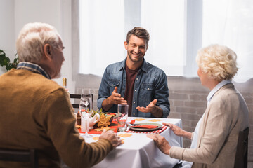 joyful man gesturing while talking to senior parents during thanksgiving dinner