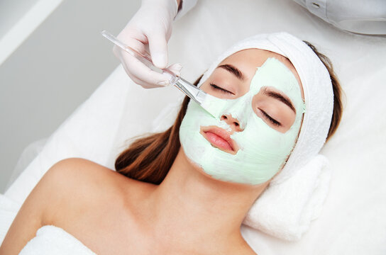 Beautiful young woman with facial mask in beauty salon. Girl getting beauty treatment facial care by beautician