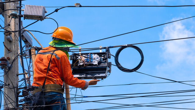 Rear view of technician on wooden ladder is installing fiber optic cable and internet splitter box on electric pole against blue sky background