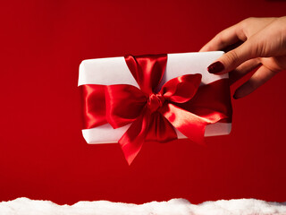 Christmas present. Winter holidays surprise. Female hand holding wrapped white gift box with ribbon bow isolated on red festive background with snow pile. Happy New year.