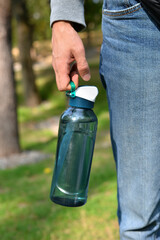 Man's hand holds blue plastic reusable sports water bottle to go, on a nature background. Concept...