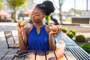 Healthy food concept. Close-up of beautiful African American woman choose eating salad and burger in outdoors cafe