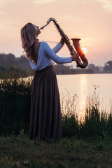 silhouette of a young beautiful girl playing the saxophone at sunrise by the river, a woman in a...