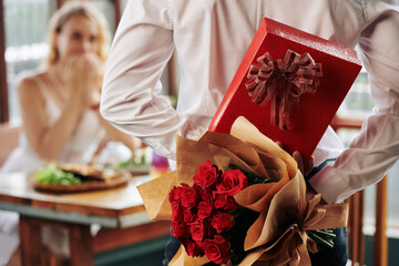 Man hiding bouquet of roses and giftbox behind his back as birthday present for girlfriend