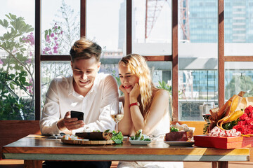 Cheerful young woman enjoying dinner in cafe and watching funny videos on smartphone