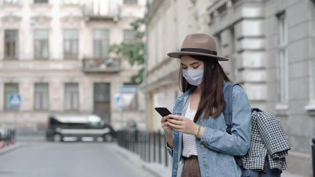 Tourist young woman wearing mask on vacation in old europe city. Portrait of beautiful girl in hat and with backpack. Using smartphone in travel