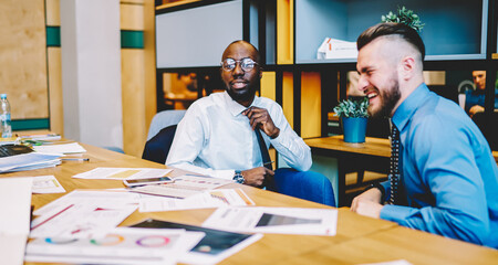 African American and Caucasian entrepreneurs in formal wear discussing business strategy for developing common project, cheerful financial directors talking about work plan analyzing paper statistics