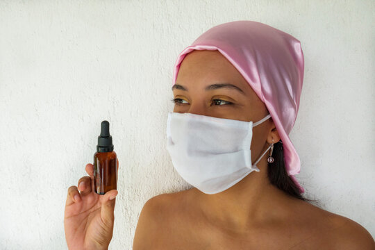 Woman with facial mask and pink scarf holding cosmetic oil for facial skin care