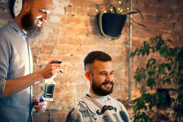 Hairdresser using hair spray on client hair at barber shop. Good looking man visiting barber shop. 
