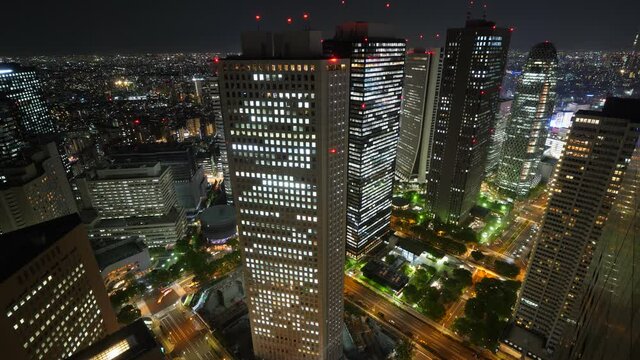 Several tall office towers with glowing lights in window, night time, cars rush on streets between buildings. Aerial time lapse shot of Asian metropolis business district, scenic cityscape