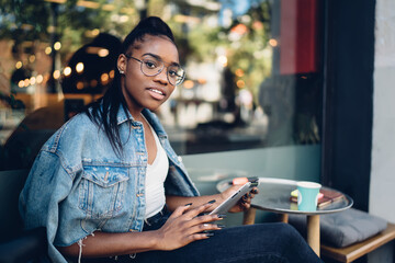 Positive trendy dressed woman in spectacles looking at camera using modern digital tablet connected to 4G internet, smiling female 20s african american looking at camera checking notifications