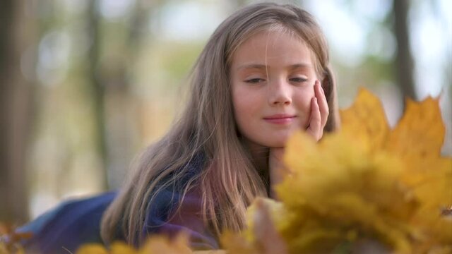 Close up portrait of a cute little girl with long blond hair lies on fallen leaves in an autumn park. Childhood Dream.