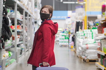 A woman with a suitcase on wheels, standing in a big store. Turns around and smiles. On the face of the woman put on a black, protective mask. Shopping and traveling during a virus and pandemic.