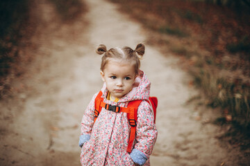 little pretty girl dressed in a pink jacket and backpack looks serious looking while walking in the...